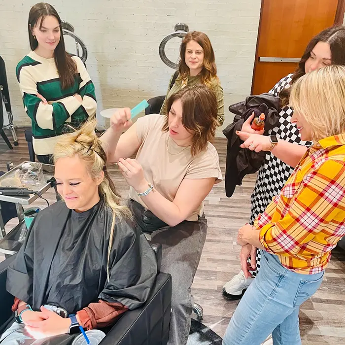 A woman gets her hair styled in a salon chair while four other women observe. The stylist works on her hair with a focused expression. The scene appears to be in a modern salon setting.