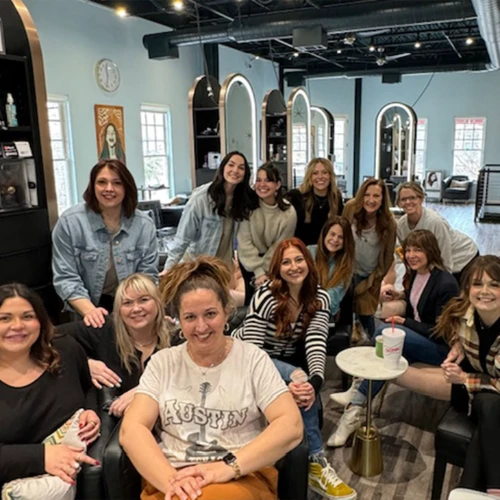 Five women are seated indoors, smiling and laughing. They are in close proximity, with shelves of various products visible behind them.